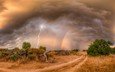 грунтовая дорога проходящая через поле на фон, грозового неба со сверкающими молниями. радуг, дождевыми потоками по линии горизонта